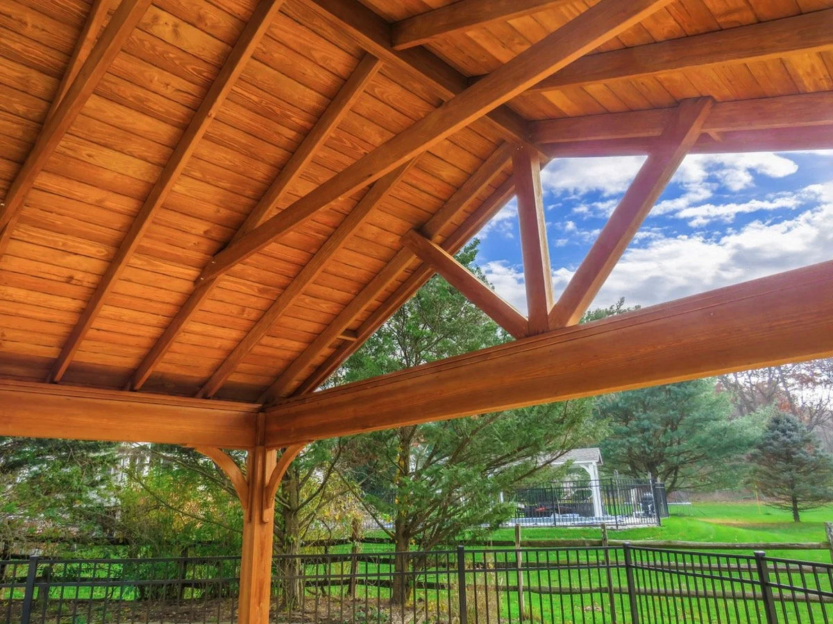 Creative Gazebos Keystone A-Frame Wood Pavilion: Close-up view of a wooden patio or porch ceiling with exposed beams, overlooking a backyard with trees, a gazebo, a black metal fence, and a partly cloudy blue sky. Manufacturer family example; size, finishes and optional features vary.