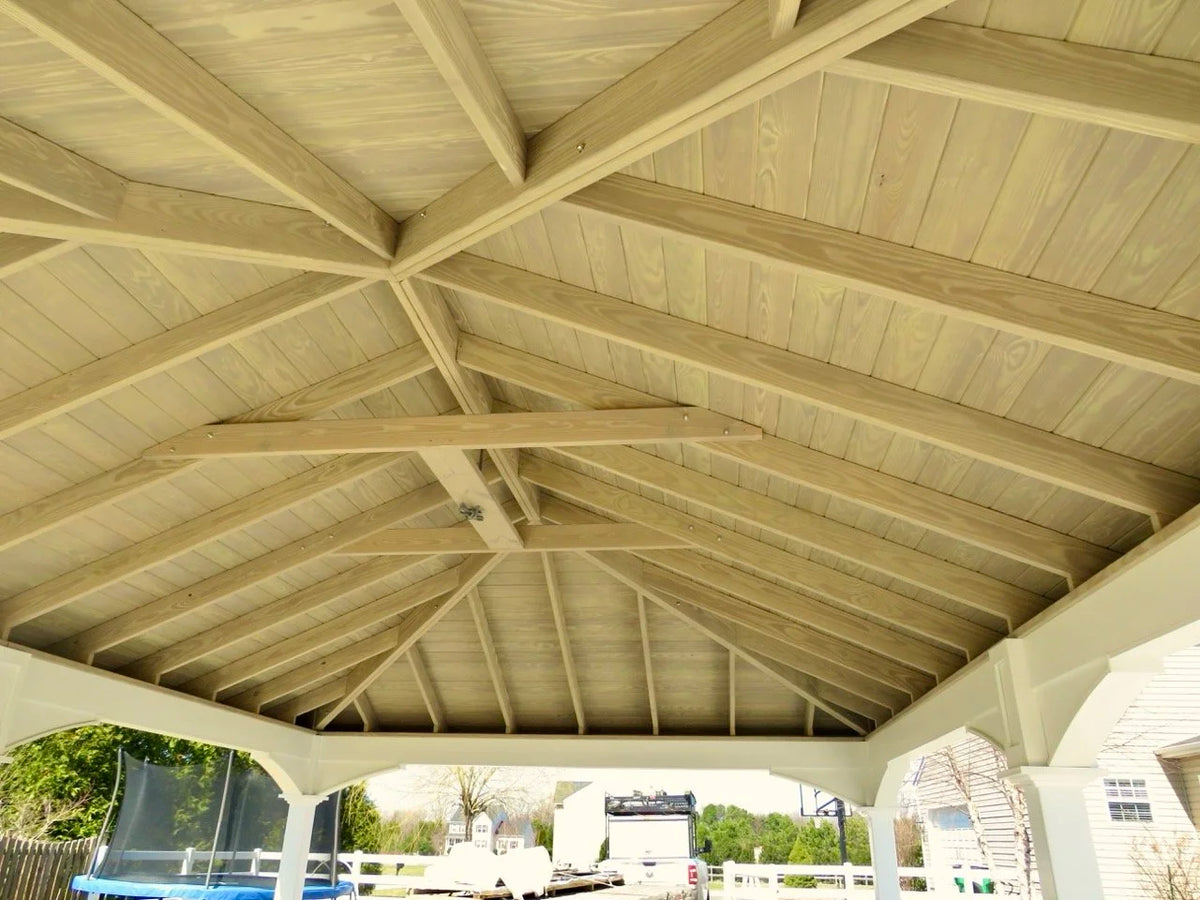 Creative Gazebos Manor Vinyl Pavilion: View of a wooden roof structure with exposed beams and a ceiling, open on the sides, with outdoor scenery including a trampoline and a house in the background.. Manufacturer family example; size, finishes and optional features vary.