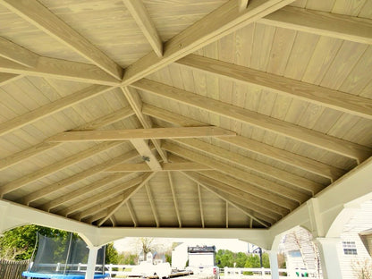 Creative Gazebos Manor Vinyl Pavilion: View of a wooden roof structure with exposed beams and a ceiling, open on the sides, with outdoor scenery including a trampoline and a house in the background.. Manufacturer family example; size, finishes and optional features vary.